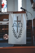 A podium with a hanging cloth featuring a religious symbol made up of a cross, the Greek letters Alpha and Omega, and a laurel wreath. A microphone is positioned next to the podium. The setting is likely within a church, indicated by the dimly lit interior and stained glass decor in the background.