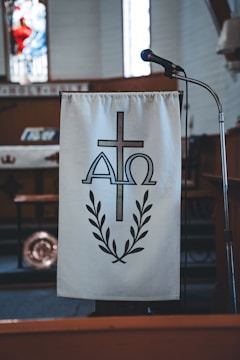 A podium with a hanging cloth featuring a religious symbol made up of a cross, the Greek letters Alpha and Omega, and a laurel wreath. A microphone is positioned next to the podium. The setting is likely within a church, indicated by the dimly lit interior and stained glass decor in the background.