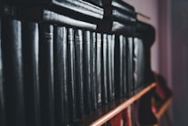 A row of dark, leather-bound books neatly lined up on a wooden shelf. The books exhibit a classic and aged appearance, suggesting they may contain collections of literature or encyclopedias. The lighting is dim, creating a moody atmosphere.