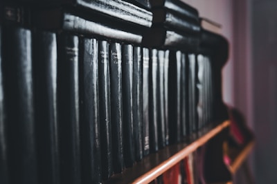 A row of dark, leather-bound books neatly lined up on a wooden shelf. The books exhibit a classic and aged appearance, suggesting they may contain collections of literature or encyclopedias. The lighting is dim, creating a moody atmosphere.