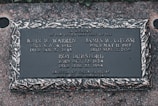 A memorial plaque dedicated to individuals who were lost at sea, featuring engraved names, birth and death dates, and a decorative border. The plaque is set against a textured stone surface.