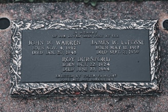 A memorial plaque dedicated to individuals who were lost at sea, featuring engraved names, birth and death dates, and a decorative border. The plaque is set against a textured stone surface.