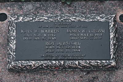 A memorial plaque dedicated to individuals who were lost at sea, featuring engraved names, birth and death dates, and a decorative border. The plaque is set against a textured stone surface.
