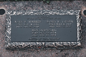 A memorial plaque dedicated to individuals who were lost at sea, featuring engraved names, birth and death dates, and a decorative border. The plaque is set against a textured stone surface.