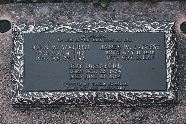 A memorial plaque dedicated to individuals who were lost at sea, featuring engraved names, birth and death dates, and a decorative border. The plaque is set against a textured stone surface.