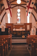 A church interior featuring wooden pews facing an altar with religious symbols and banners. The ceiling has a wooden arch design, and there are three bright overhead lights above a stained glass window depicting a religious scene.