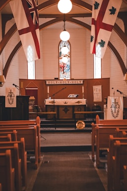 A church interior featuring wooden pews facing an altar with religious symbols and banners. The ceiling has a wooden arch design, and there are three bright overhead lights above a stained glass window depicting a religious scene.