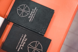 Two black books titled 'The Book of Alternative Services of The Anglican Church of Canada' are placed on an orange surface. The cover features a simple cross emblem.