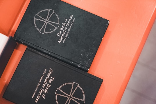 Two black books titled 'The Book of Alternative Services of The Anglican Church of Canada' are placed on an orange surface. The cover features a simple cross emblem.
