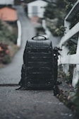 A large black backpack is positioned on a narrow path with a blurred background of houses and greenery. The backpack seems rugged and well-equipped, possibly for travel or photography. To the side, an object resembling a tripod or monopod is attached.