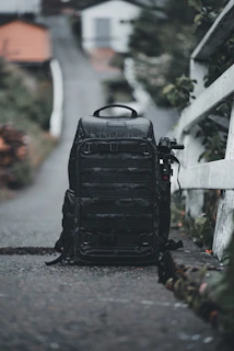 A large black backpack is positioned on a narrow path with a blurred background of houses and greenery. The backpack seems rugged and well-equipped, possibly for travel or photography. To the side, an object resembling a tripod or monopod is attached.