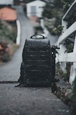 A large black backpack is positioned on a narrow path with a blurred background of houses and greenery. The backpack seems rugged and well-equipped, possibly for travel or photography. To the side, an object resembling a tripod or monopod is attached.