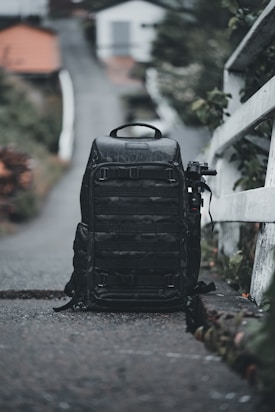 A large black backpack is positioned on a narrow path with a blurred background of houses and greenery. The backpack seems rugged and well-equipped, possibly for travel or photography. To the side, an object resembling a tripod or monopod is attached.