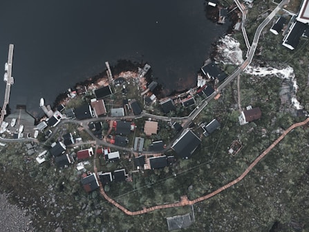 An aerial view of a small coastal village with various buildings scattered around a winding road. The village is situated next to a body of water with several docks and boats. The landscape around the village is green and rocky, with some trees and pathways visible.