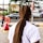 An individual with long, tightly braided hair stands on a city sidewalk. The person is wearing a white t-shirt, and the scene is set near a busy road with passing cars. In the background, several signs and advertisements are visible, including one for KFC. Trees and other urban elements are part of the backdrop.