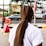 An individual with long, tightly braided hair stands on a city sidewalk. The person is wearing a white t-shirt, and the scene is set near a busy road with passing cars. In the background, several signs and advertisements are visible, including one for KFC. Trees and other urban elements are part of the backdrop.