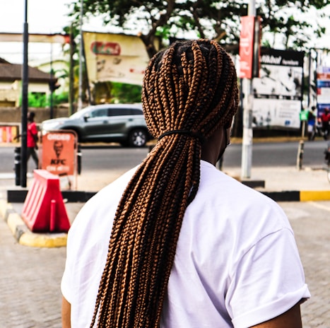 An individual with long, tightly braided hair stands on a city sidewalk. The person is wearing a white t-shirt, and the scene is set near a busy road with passing cars. In the background, several signs and advertisements are visible, including one for KFC. Trees and other urban elements are part of the backdrop.