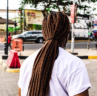 An individual with long, tightly braided hair stands on a city sidewalk. The person is wearing a white t-shirt, and the scene is set near a busy road with passing cars. In the background, several signs and advertisements are visible, including one for KFC. Trees and other urban elements are part of the backdrop.