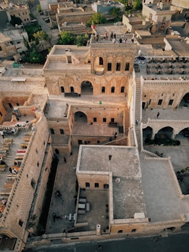 An aerial view of a historical stone building complex with multiple levels and arched windows. People are standing on the rooftop and in the courtyard area. The surrounding neighborhood features a mix of traditional and modern buildings, with some greenery visible. The lighting suggests late afternoon or early evening.