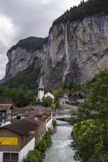A picturesque village nestled in a lush mountainous landscape, featuring a dramatic waterfall cascading down a steep cliff. Traditional wooden houses with sloped roofs line the river, which flows through the valley. A prominent church with a tall spire stands among the buildings, surrounded by dense green trees.
