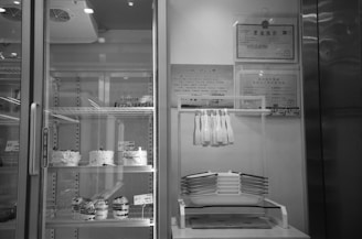A display case features several cakes and desserts on multiple shelves. To the right, a rack holds white plastic spatulas above a stack of white trays. Certificates are framed and mounted on the wall above the rack.