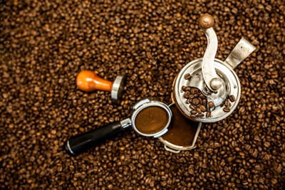 A metal coffee grinder and a tamper are placed on a bed of roasted coffee beans. Some coffee beans are visible inside and around the grinder, and ground coffee is present in the grinder drawer and the coffee portafilter.