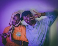 A man and woman standing closely together against a softly lit background with a purple hue. The woman is wearing a patterned headscarf and an orange shirt, while the man is dressed in a pastel shirt and a dark cap, flashing a peace sign. Both individuals are smiling, creating a relaxed and joyful atmosphere.