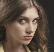 A close-up portrait of a woman with long, wavy brown hair. She has a serious and intense expression, gazing directly at the camera. Her makeup is subtle, emphasizing her eyes and lips, with a soft light illuminating her face against a dark background.