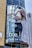 A large billboard on a building displays an advertisement featuring a woman in a white outfit posing inside a blue vintage car. The background shows a modern glass building and part of another structure under construction. The text on the billboard reads 'bella' and 'SOFT ON YOU, SOFTER'.