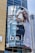 A large billboard on a building displays an advertisement featuring a woman in a white outfit posing inside a blue vintage car. The background shows a modern glass building and part of another structure under construction. The text on the billboard reads 'bella' and 'SOFT ON YOU, SOFTER'.
