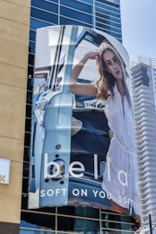 A large billboard on a building displays an advertisement featuring a woman in a white outfit posing inside a blue vintage car. The background shows a modern glass building and part of another structure under construction. The text on the billboard reads 'bella' and 'SOFT ON YOU, SOFTER'.