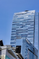 A tall, modern skyscraper with a sleek, glass exterior and a reflective surface under a clear blue sky. The building features the logo of 'The Ritz-Carlton' at the top. In the foreground, part of a traffic light and another building with a contemporary design can be seen.