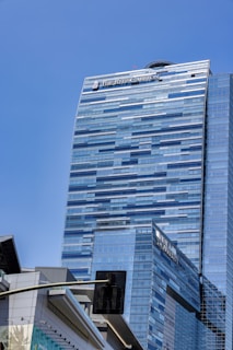 A tall, modern skyscraper with a sleek, glass exterior and a reflective surface under a clear blue sky. The building features the logo of 'The Ritz-Carlton' at the top. In the foreground, part of a traffic light and another building with a contemporary design can be seen.