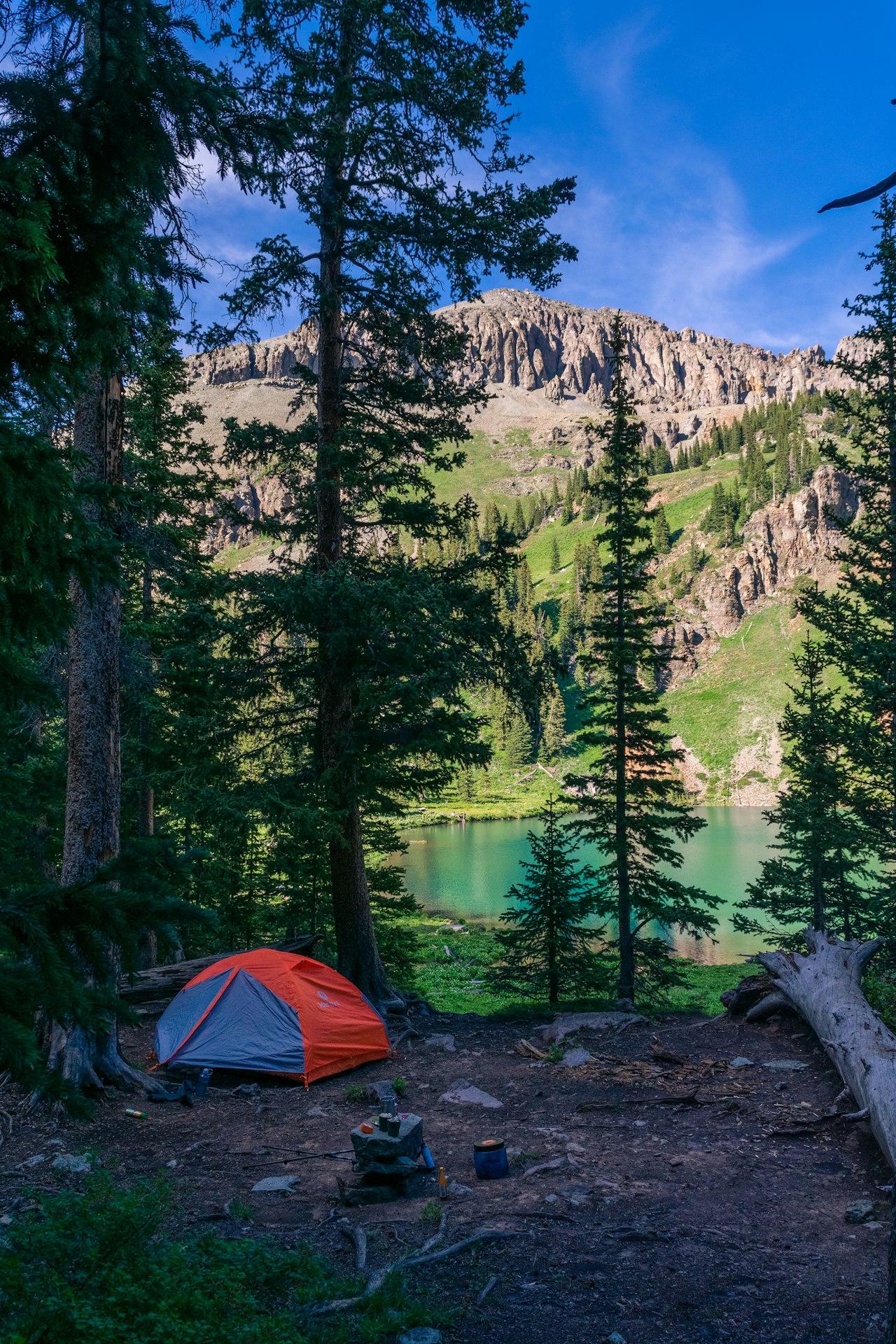 A tent pitched up in the woods next to a lake. There are trees and mountains in the background.