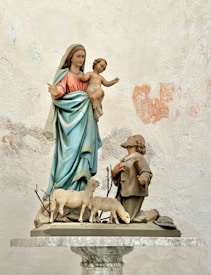 A religious statue depicts a woman holding a child, standing on a pedestal alongside a kneeling man. Surrounding them are several sheep. The woman is draped in robes of blue and red, while the child is nude. The man appears to be in a posture of adoration or prayer. The statue is set against a textured, weathered wall.