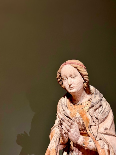 A wooden sculpture of a figure with a serene expression, dressed in detailed drapery. The figure is in a praying posture, with hands clasped together and an intricate head covering.