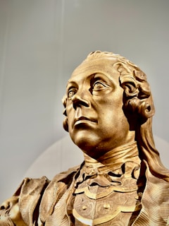 A detailed, ornate sculpture of a person's upper body, showcasing a classical style with intricate designs on the clothing and a solemn, dignified expression. The sculpture appears to have a metallic, gold-like finish, highlighting the craftsmanship involved.