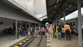 A busy train station platform with numerous people waiting and walking around. A train is stationary on the right side, while passengers, including a security officer in a bright orange vest, are either boarding or disembarking. The platform is covered with a metal roof and the atmosphere is lively with visible social interactions. The scene is bustling with various people carrying bags and the platform edge marked with yellow and black stripes.