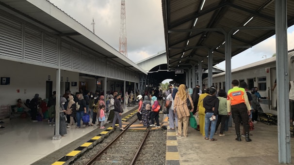 A busy train station platform with numerous people waiting and walking around. A train is stationary on the right side, while passengers, including a security officer in a bright orange vest, are either boarding or disembarking. The platform is covered with a metal roof and the atmosphere is lively with visible social interactions. The scene is bustling with various people carrying bags and the platform edge marked with yellow and black stripes.