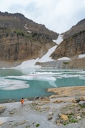 A person in a bright orange jacket stands near a rocky shoreline, overlooking a tranquil glacial lake with floating icebergs. Steep, rugged mountains rise in the background with patches of snow still visible.