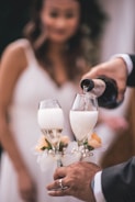 A person in formal attire pours champagne into two decorated glasses, which have floral accents. In the background, a woman in a white dress is blurred, suggesting a wedding or celebration.
