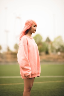 A person with pink hair is standing outdoors wearing an oversized pink sweatshirt. The background is a blurred view of trees and an outdoor sports field.