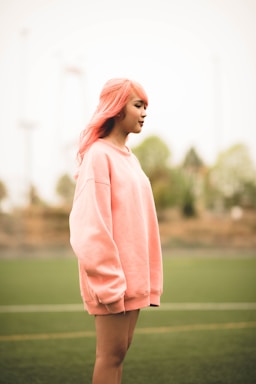A person with pink hair is standing outdoors wearing an oversized pink sweatshirt. The background is a blurred view of trees and an outdoor sports field.