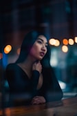 A person gazes thoughtfully into the distance while leaning against a wooden surface. Warm, blurred lights in the background add a sense of depth and ambiance to the scene. The person's expression suggests contemplation or introspection.