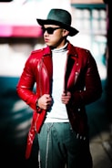 A person wearing a stylish red leather jacket over a white turtleneck, accessorized with a grey wide-brimmed hat and dark sunglasses. The individual has hands adjusting the jacket, and they are standing on a sunlit street with blurred buildings in the background.