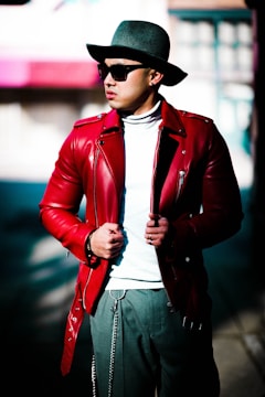 A person wearing a stylish red leather jacket over a white turtleneck, accessorized with a grey wide-brimmed hat and dark sunglasses. The individual has hands adjusting the jacket, and they are standing on a sunlit street with blurred buildings in the background.