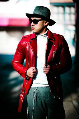 A person wearing a stylish red leather jacket over a white turtleneck, accessorized with a grey wide-brimmed hat and dark sunglasses. The individual has hands adjusting the jacket, and they are standing on a sunlit street with blurred buildings in the background.