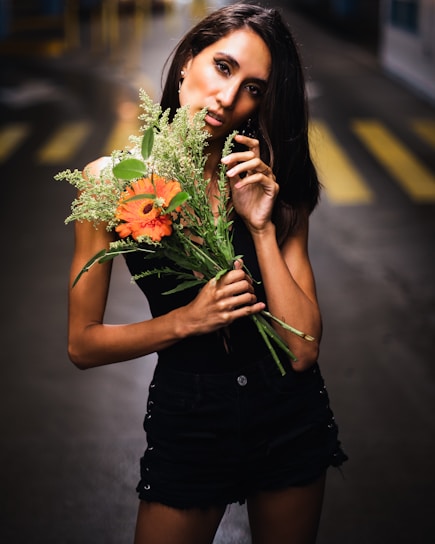 A woman stands in an alley holding a bouquet of orange and green flowers. She has long dark hair and is wearing a black sleeveless top with black shorts. The background is blurred, featuring yellow pedestrian crossing lines.