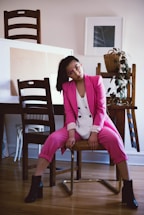 A woman wearing a pink suit and white blouse is seated on a chair in a casual pose. She has dark hair pulled back in a ponytail and is wearing black ankle boots. The setting appears to be a home or studio with wooden furniture, including chairs and a table. There are framed pictures on the wall and a plant in a woven basket on the table.