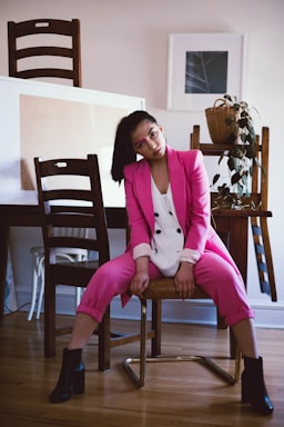 A woman wearing a pink suit and white blouse is seated on a chair in a casual pose. She has dark hair pulled back in a ponytail and is wearing black ankle boots. The setting appears to be a home or studio with wooden furniture, including chairs and a table. There are framed pictures on the wall and a plant in a woven basket on the table.